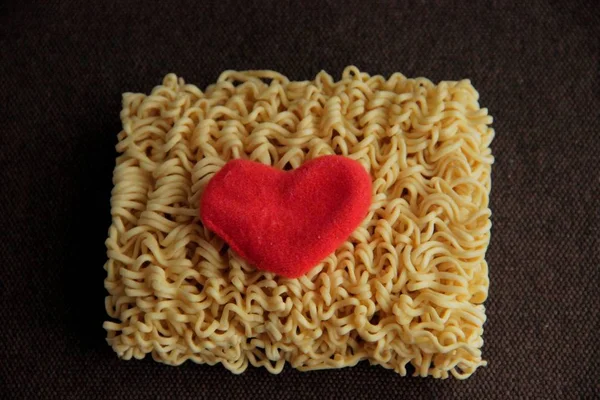 dry yellow noodles and heart made of red fleece fabric on brown background close up and blurred.