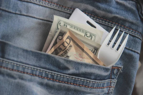 Small american bills, card key and fork in a pocket of blue jeans of office employee hurrying on a lunch break close up. Concept of fast food in beat big city. blur