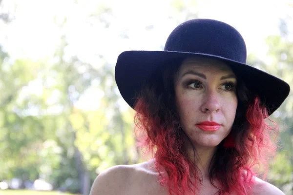 Mujer europea con lápiz labial rojo en un día soleado. Mujeres en un sombrero negro con ala, con el pelo rizado rojo . — Foto de Stock