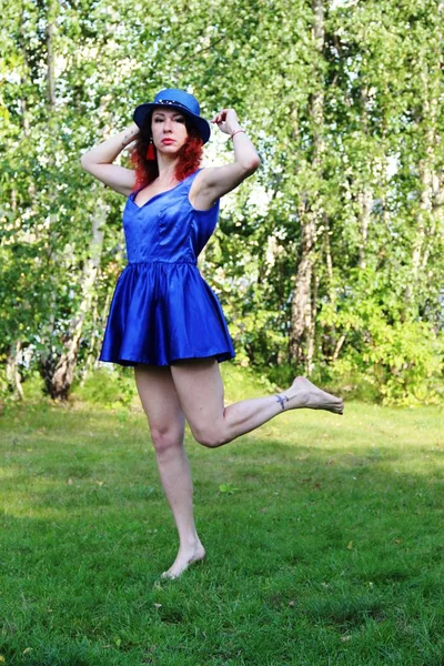 Woman on the lawn with red curly hair in a blue hat and short blue mini dress. Holding a hat. stands on one leg. — Stock Photo, Image