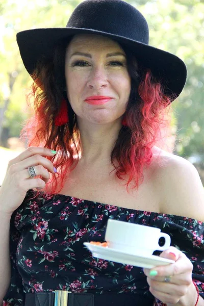 Mujer con rizos rojos de pelo, vestida con un vestido negro y un sombrero negro, con una taza de té en la mano, vertical — Foto de Stock