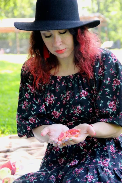 siyah şapkalı kırmızı saçlı, kıvırcık saçlı bir kadın, eşleşen bir elbise ve kırmızı püskül küpe avuçiçlerinde marmelat şekerleri bakar