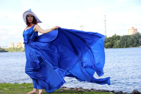 Portrait in full growth on the banks of the river, in a windy warm summer day. woman in a gorgeous long blue dress — Stock Photo, Image