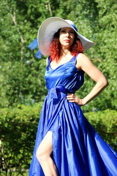 Une dame en robe bleue et un chapeau blanc sur une pelouse verte. une femme au chapeau et une robe longue avec un train sur la jupe dans le parc, verticale — Photo