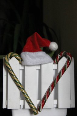 İki şeker kamışı ve beyaz dekoratif çitin üzerinde ponponu olan kırmızı bir Noel şapkası, dikey, kahverengi arka plan.