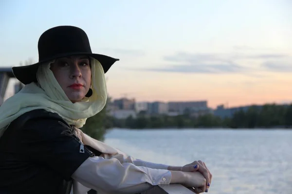 Una mujer con una chaqueta negra, un sombrero negro sobre un pañuelo beige, sola junto al río. después del autoaislamiento, retrato sin guantes y máscaras — Foto de Stock