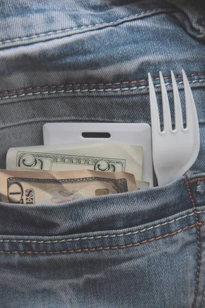 Small American bills, a card key, and a plastic fork in the pocket of the blue jeans of an employee rushing to a lunch break. Fast food concept in a big city.