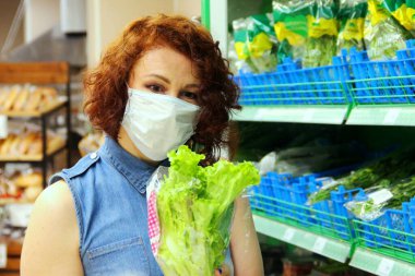 Tıbbi maskeli esmer dükkandan yeşil salata alır. Sebzelerle dolu rafın yanındaki salataya bak, seçici odaklı.. 