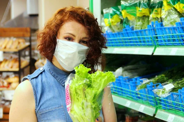 Brunette in medical mask buys green salad in store. looks at the salad next to the shelf with vegetables, selective focus.