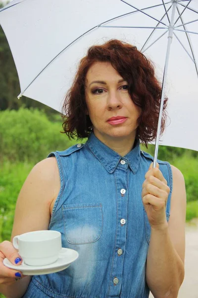 Morena Mujer en vestido de mezclilla con una taza de café o té bajo un paraguas blanco en la naturaleza. llovizna — Foto de Stock
