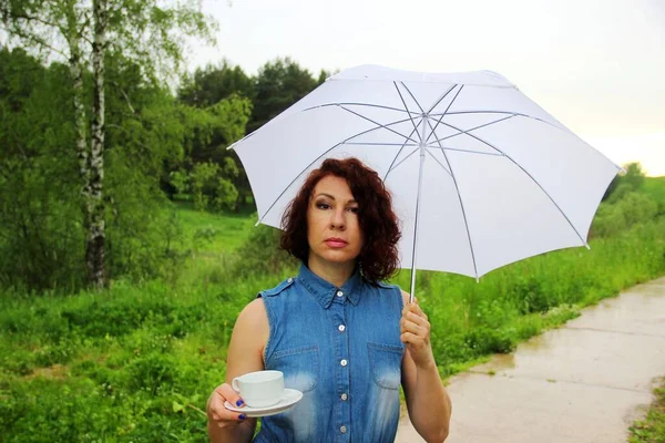 Encantador morena en vestido de mezclilla con una taza de café bajo un paraguas blanco en la naturaleza. espolvorear — Foto de Stock