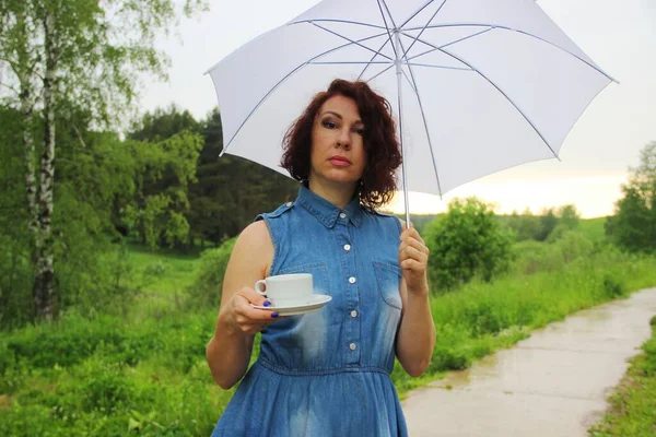 Encantador morena en vestido de mezclilla con una taza de té bajo un paraguas blanco en la naturaleza. llovizna — Foto de Stock