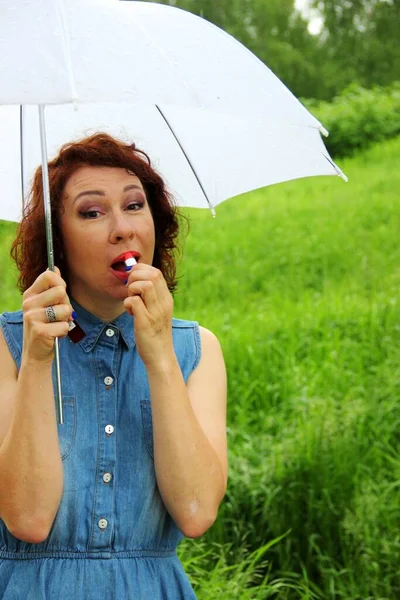 Attractive brunette in a denim dress with a white umbrella in one hand touches up her lips with red lipstick. — Stock Photo, Image