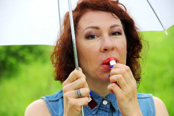Atractiva morena en vestido de mezclilla con paraguas blanco en una mano, pinta labios con lápiz labial rojo — Foto de Stock