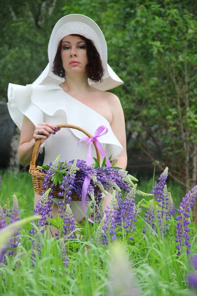 Woman in a white hat with a basket of lilac lupines, vertical photo. A brunette in a white dress and a white hat collects lupine flowers. — Stock Photo, Image