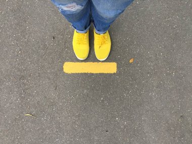 Sarı çizme ve mavi kot pantolon asfalttaki sarı dur çizgisinde, yukarıdan görünüyor. İşaretlemeyi durdur, sosyal uzaklık. 