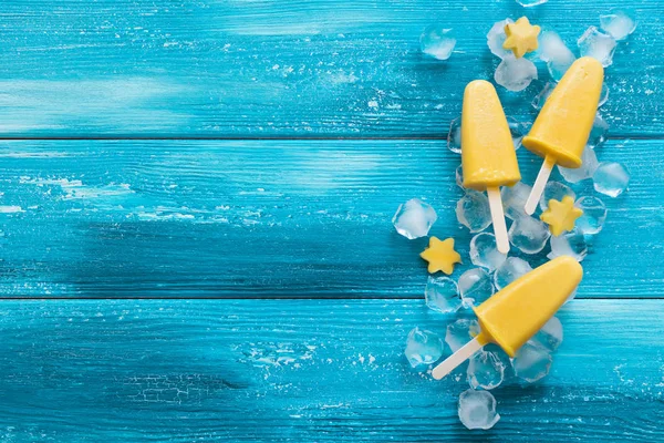 Yellow mango ice cream on a stick on a blue wooden background. Fruit frozen dessert. Copy space. View from above. Flat layout.