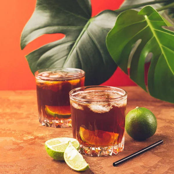 Een drankje van rum of Cola ijsblokjes en sappige kalk in glazen bekers tegen een achtergrond van tropische groene bladeren. — Stockfoto