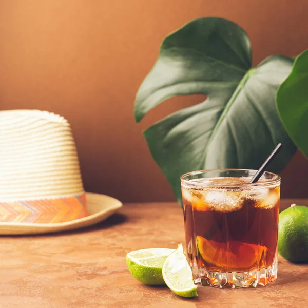 Koude Iced Tea in een glas op de achtergrond van de bladeren van tropische plant. Modieuze koraal achtergrond en zomer hoed. — Stockfoto