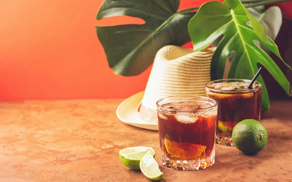 Té helado frío en un vaso en el fondo de las hojas de la planta tropical. Fondo de coral de moda y sombrero de verano . — Foto de Stock