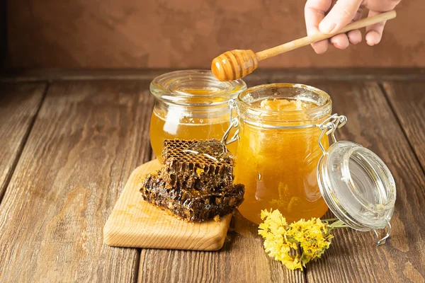 A hand is holding awooden spoon which honey flows. Banks of fresh golden flower honey on a wooden background the table.