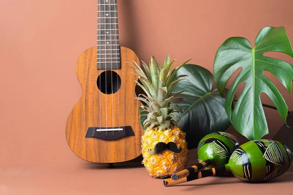 Funny dressed-up pineapple with black mustache. Maracas Guitar on the background of large green leaves of the Monstera.