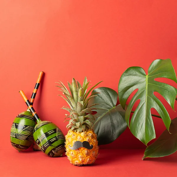 Ripe pineapple with black mustache maracas big leaves of monstera red bright background. The concept of a musical party.