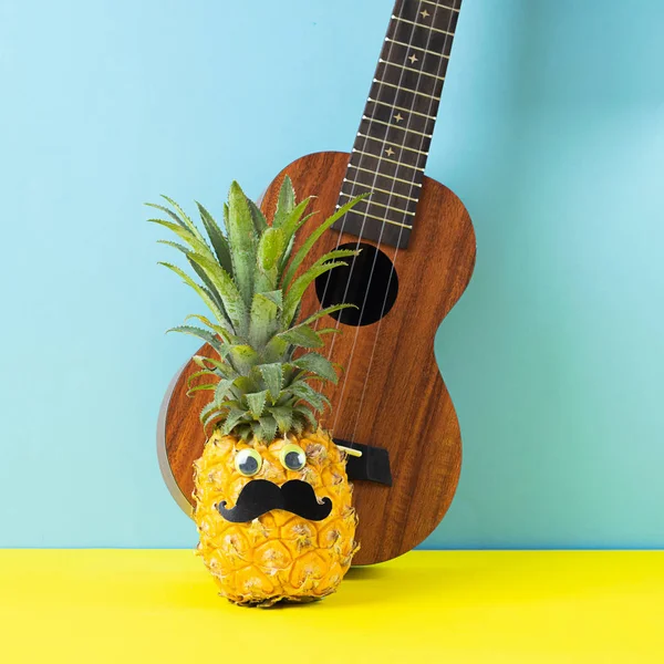 Rijpe ananas met zwarte snor ogen gitaar op een geel blauwe achtergrond. Grappig gezicht van voedsel. Het concept van de zomer — Stockfoto