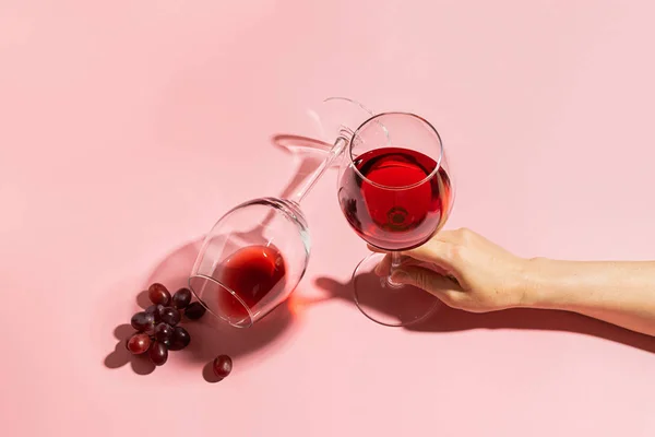 Vrouwelijke hand houden van een glas met rode wijn en tros druiven op een zachte roze achtergrond. Selectieve focus. Minimalisme. — Stockfoto