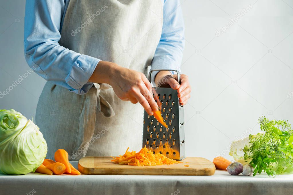 Woman grates fresh carrots for cooking salad or for salting cabbage natural background. Vegetarian food concept.