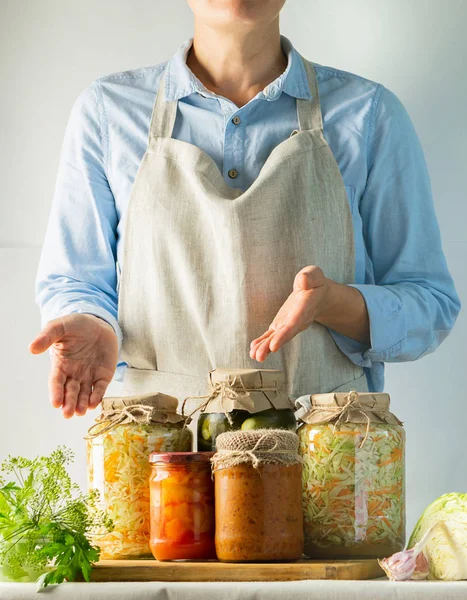 Las verduras fermentadas y enlatadas varias latas están sobre la mesa y una mujer cocina en un delantal. Reciclaje de la cosecha de otoño — Foto de Stock