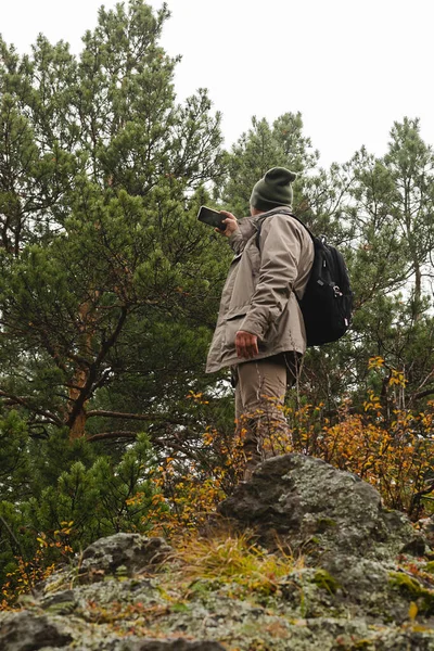 Un viajero hombre con una mochila se para en el borde de la montaña y toma una foto selfie en su teléfono móvil . —  Fotos de Stock