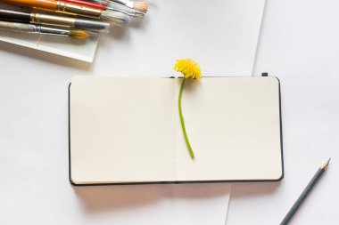 Temiz, beyaz bir defter boya fırçalarıyla dolu bir masada yatıyor. Düz yerleşim yeri tasarım metni. Sanat yaratıcılığı kavramı