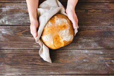 Bir somun taze pişmiş ekmeği altın hamuruyla tutan eller. Ev yapımı mayasız hamur işleri ahşap arka plan.
