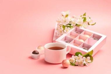 Şenlikli zarif bir kutuda çeşitli renkte çikolatalar. Pastel pembe arka plan dekorasyon çiçeklerinde bir fincan çay paketliyorlar..