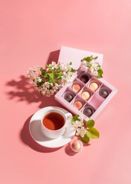 Şenlikli zarif bir kutuda çeşitli renkte çikolatalar. Pastel pembe arka plan dekorasyon çiçeklerinde bir fincan çay paketliyorlar..