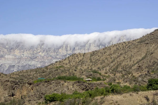 La gran montaña se encuentra alta en el cielo azul; rodeada de verdes pendientes y está parcialmente cubierta por las nubes bajas que yacen . — Foto de Stock