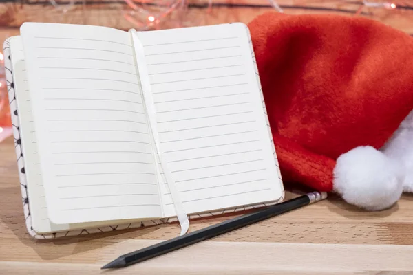 Not defteri, kırmızı Noel şapkası ve kalem arka plandaki ahşap masada Noel çelengi.