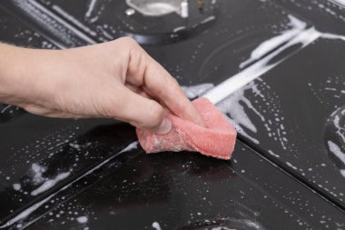 Kadın eli gaz sobasını, cam seramik yüzeyi yıkıyor. Kırmızı sünger ve beyaz köpük..