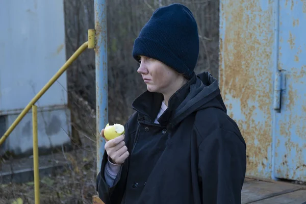 Uma sem-abrigo de casaco preto e chapéu azul está a comer uma maçã velha de latas de lixo de rua. pobreza . — Fotografia de Stock