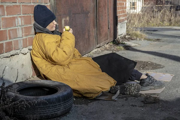 Mulher sem-teto em um amarelo velho casaco irregular e chapéu azul está sentado em papelão no pavimento e há uma velha maçã — Fotografia de Stock