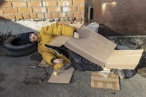 Egy hajléktalan nő sárga szakadt kabátban és kék kalapban fekszik, és kartonpapíron alszik a járdán. — Stock Fotó
