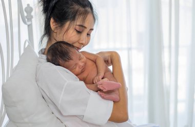 Asya beyaz gömlek anne göğsünde onu küçük uyuyan yeni doğan bebek tutar ve beyaz perde ile cam pencereler önünde beyaz yatakta oturup