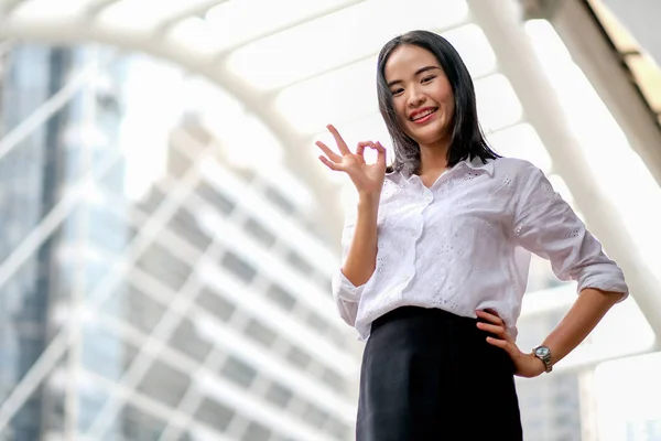 Asian beautiful business girl with white shirt act as confident and show OK sign in big city in day time.