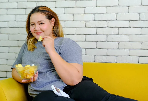 Fat girl with grey t-shirt is eating potatoes chips and sitting on yellow sofa during watch TV show in her house.