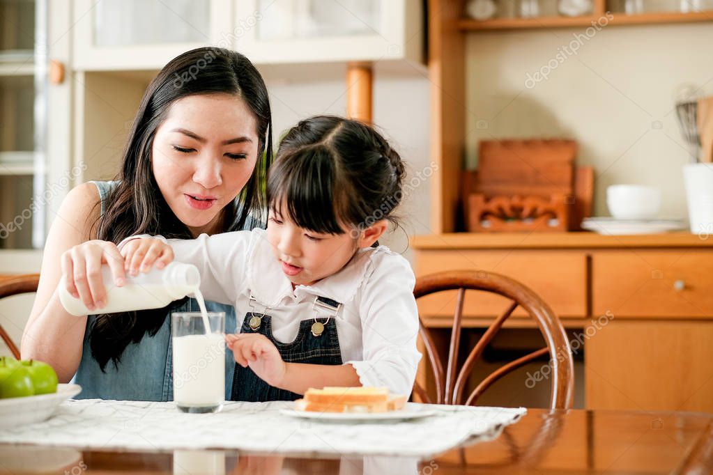 Asian mother take care her daughter pour milk to a glass on the table in house kitchen during breakfast meal. Main focus is the mother.