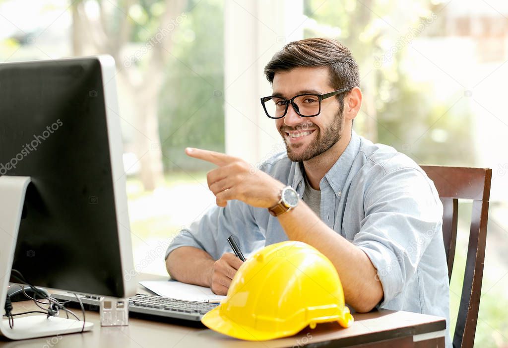 Handsome engineer man with smiling point his finger to computer monitor and sit in the office with yellow helmet on the desk and glass window.