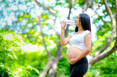 Güzel Asyalı hamile kadın gün ışığı ile yeşil bahçede açık plastik şişeden su içmek.