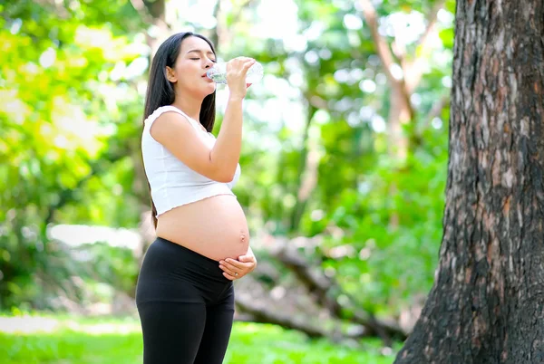 美丽的亚洲孕妇喝水从透明塑料瓶在绿色花园与天光 — 图库照片