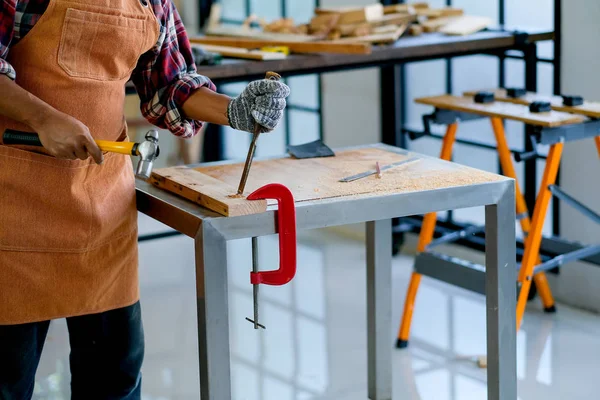 Old craftsman use hammer and chisel to work with wood and produce some product in his workplace during day time.